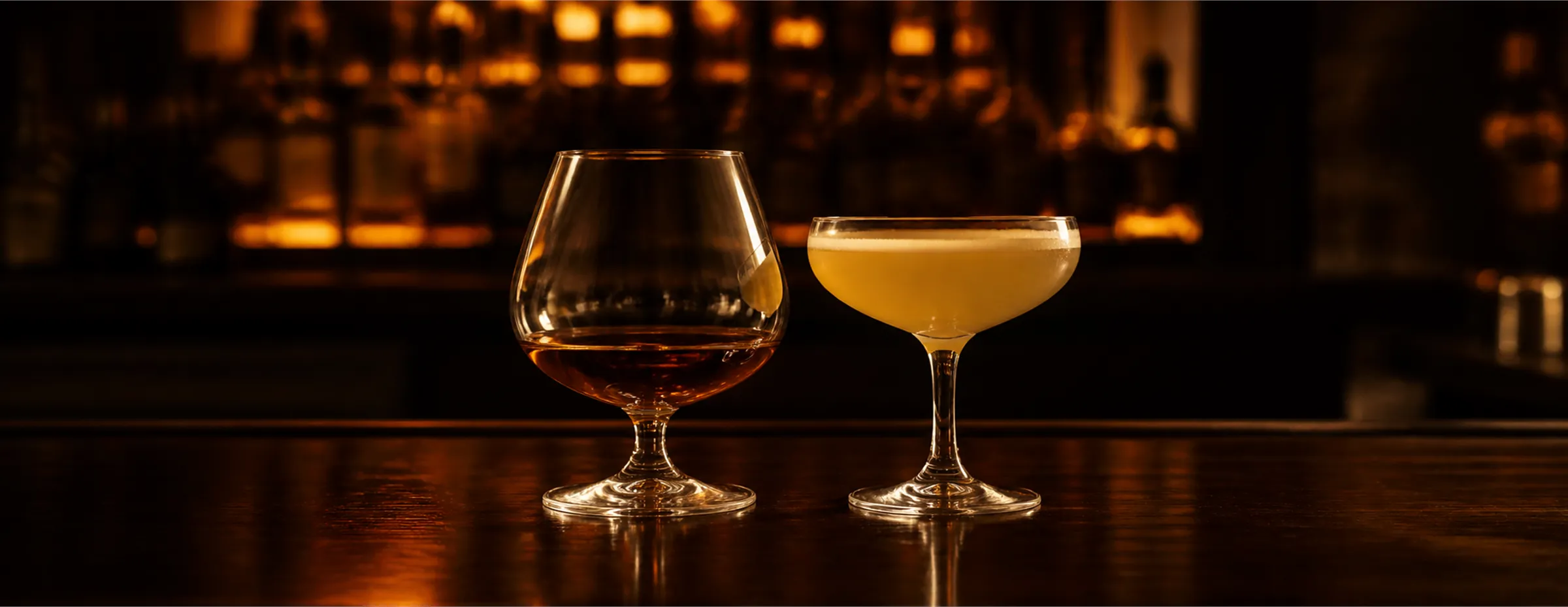 A glass of red wine beside an amber cocktail on the bar, bottles glowing behind
